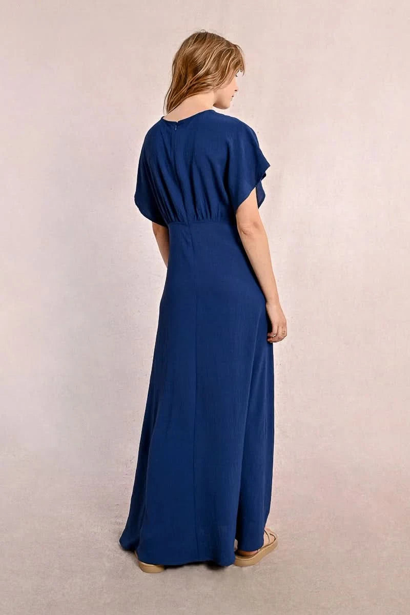 A person elegantly poses against a plain backdrop in a blue Front Pleat Long Marine Dress by Molly Bracken, characterized by its flowy design and short sleeves.