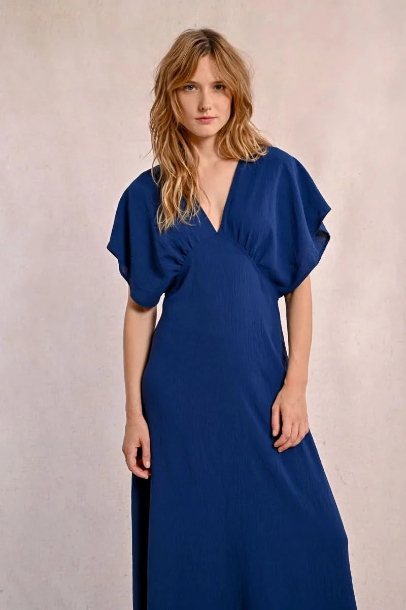 A person elegantly poses against a plain backdrop in a blue Front Pleat Long Marine Dress by Molly Bracken, characterized by its flowy design and short sleeves.