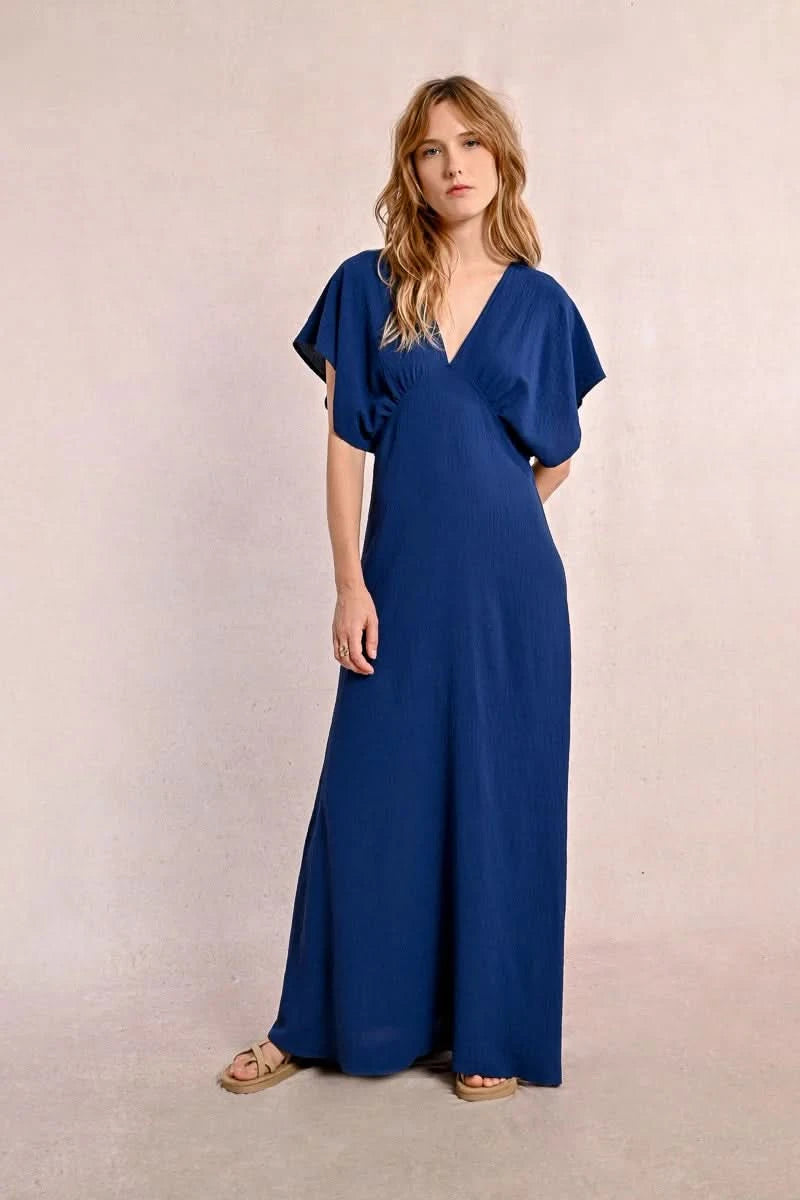 A person elegantly poses against a plain backdrop in a blue Front Pleat Long Marine Dress by Molly Bracken, characterized by its flowy design and short sleeves.