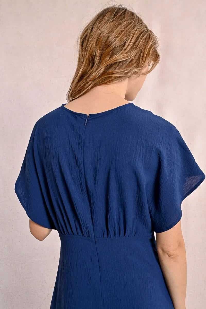 A person elegantly poses against a plain backdrop in a blue Front Pleat Long Marine Dress by Molly Bracken, characterized by its flowy design and short sleeves.