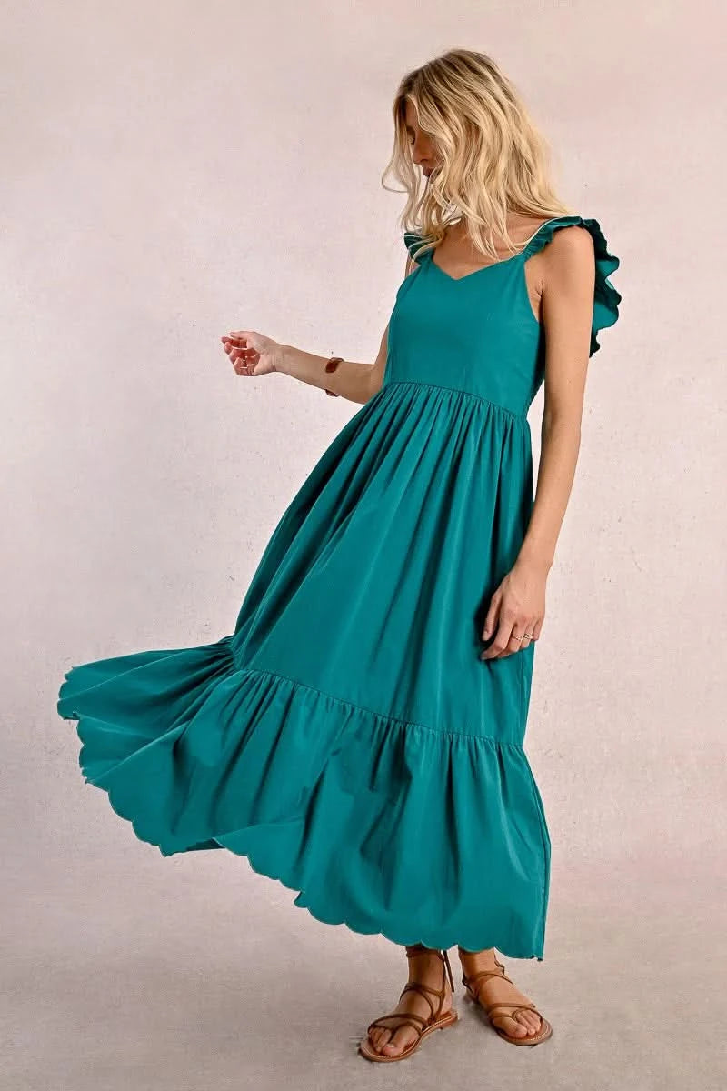 A blonde woman faces away wearing a Molly Bracken Long Ruffled Dress in teal, featuring sleeve and tier ruffles. She pairs it with brown sandals and a bracelet, set against a plain background.