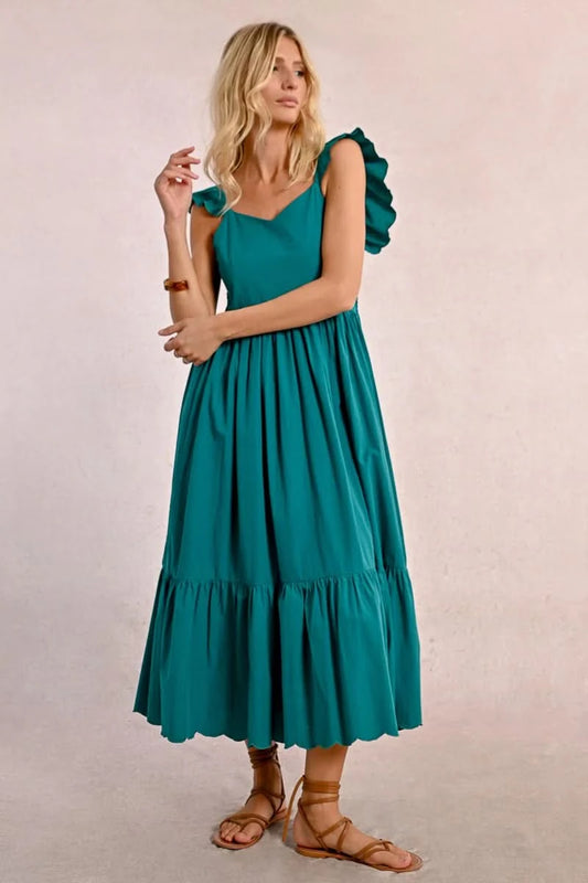 A blonde woman faces away wearing a Molly Bracken Long Ruffled Dress in teal, featuring sleeve and tier ruffles. She pairs it with brown sandals and a bracelet, set against a plain background.