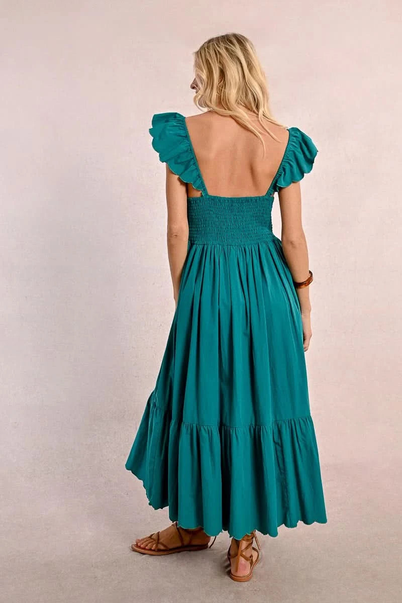 A blonde woman faces away wearing a Molly Bracken Long Ruffled Dress in teal, featuring sleeve and tier ruffles. She pairs it with brown sandals and a bracelet, set against a plain background.