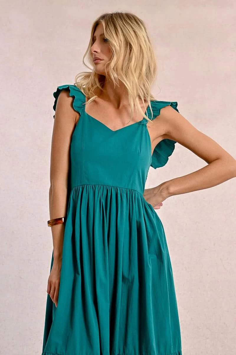 A blonde woman faces away wearing a Molly Bracken Long Ruffled Dress in teal, featuring sleeve and tier ruffles. She pairs it with brown sandals and a bracelet, set against a plain background.
