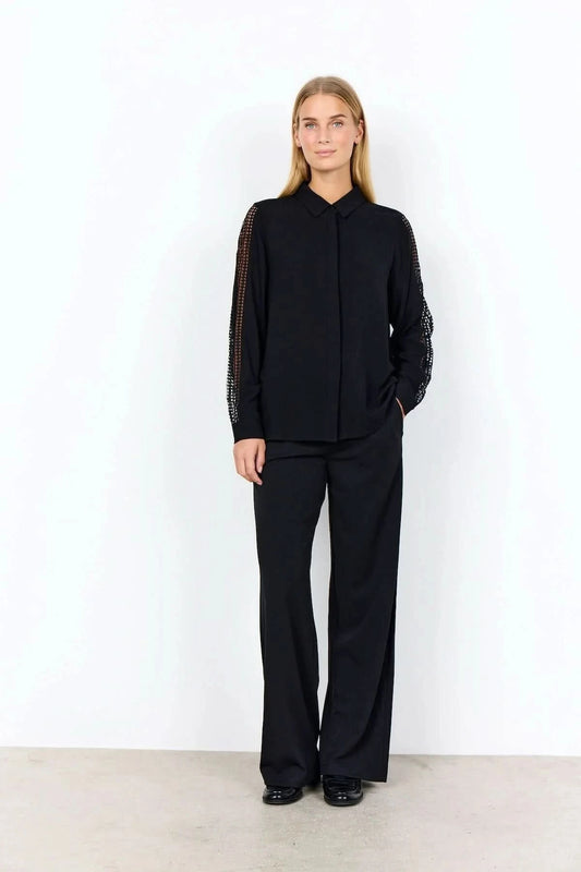 A woman stands against a plain background wearing the Soya Concepts Ladies Woven Blouse—a black, lightweight viscose blouse with lace long sleeves—paired with black pants.