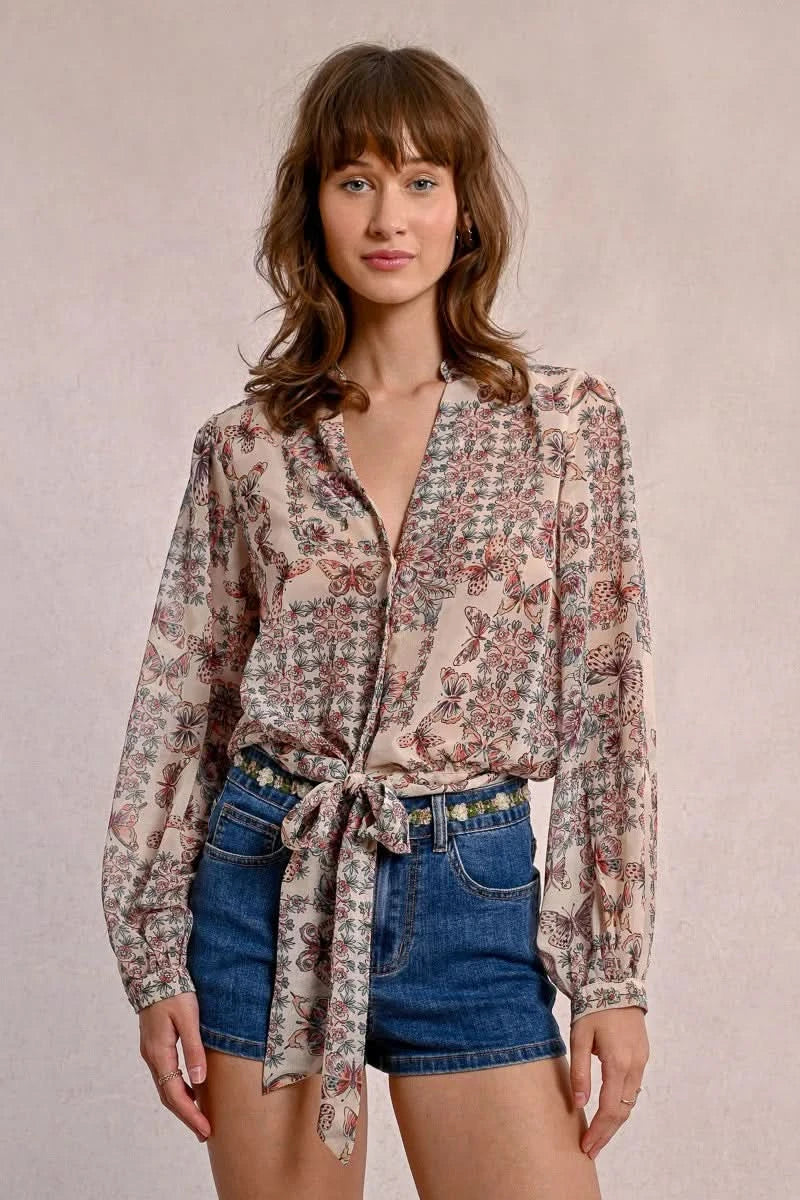 A person in a semi-transparent floral blouse and denim shorts stands against a plain background, elegantly showcasing the Front Tie Printed Blouse by Molly Bracken.