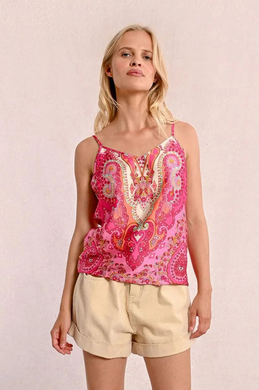 A person with long blonde hair sits on the floor against a plain background, wearing a stylish and comfortable pink patterned top from Molly Bracken called the Backless Camisole, paired with beige shorts.