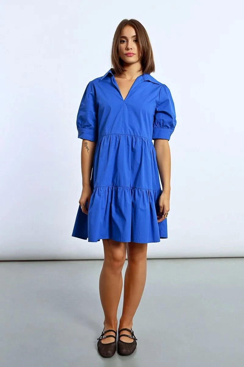 A woman stands elegantly in a Molly Bracken Ruffled Shirt Dress, tiered and blue, paired with black shoes, her hands resting by her sides against a plain background.