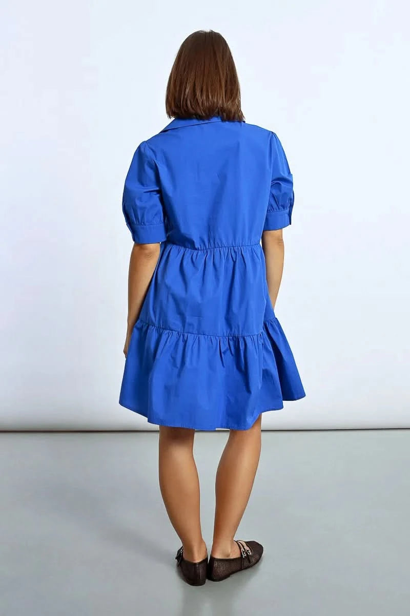 A woman stands elegantly in a Molly Bracken Ruffled Shirt Dress, tiered and blue, paired with black shoes, her hands resting by her sides against a plain background.