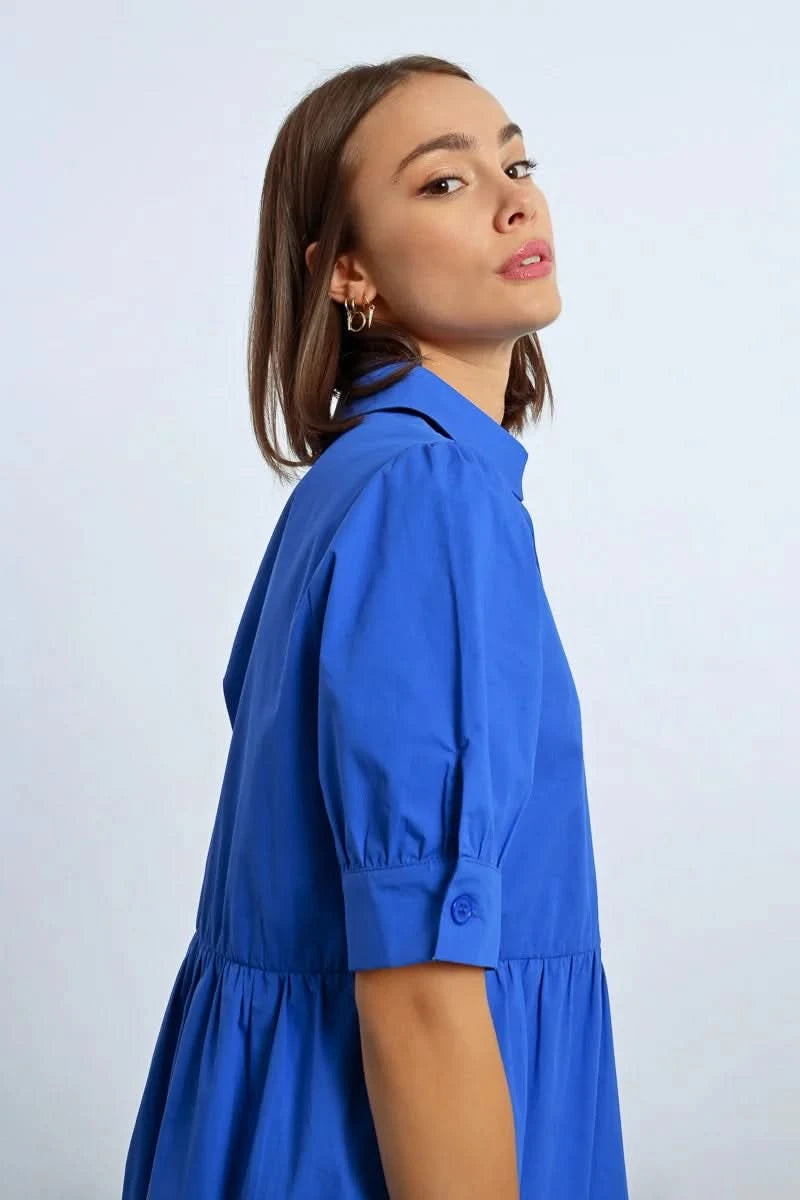 A woman stands elegantly in a Molly Bracken Ruffled Shirt Dress, tiered and blue, paired with black shoes, her hands resting by her sides against a plain background.