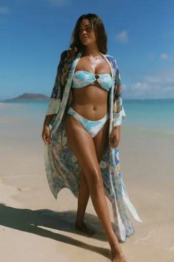 A woman in a blue and white bikini and the Tribal 3/4 SLEEVE DUSTER COVER UP strolls along a sandy beach, exuding chic vacation style with the ocean and a distant island behind her.