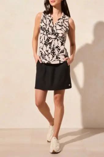 A person modeling the Tribal brand's sleeveless pebble polo top with shirttail, paired with a black skirt and white sneakers, poses against a beige background.