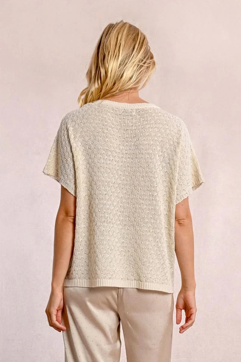 A person with long blond hair stands against a light wall, hands in pockets, wearing the Short Sleeve Knit Top by Molly Bracken and loose pants.