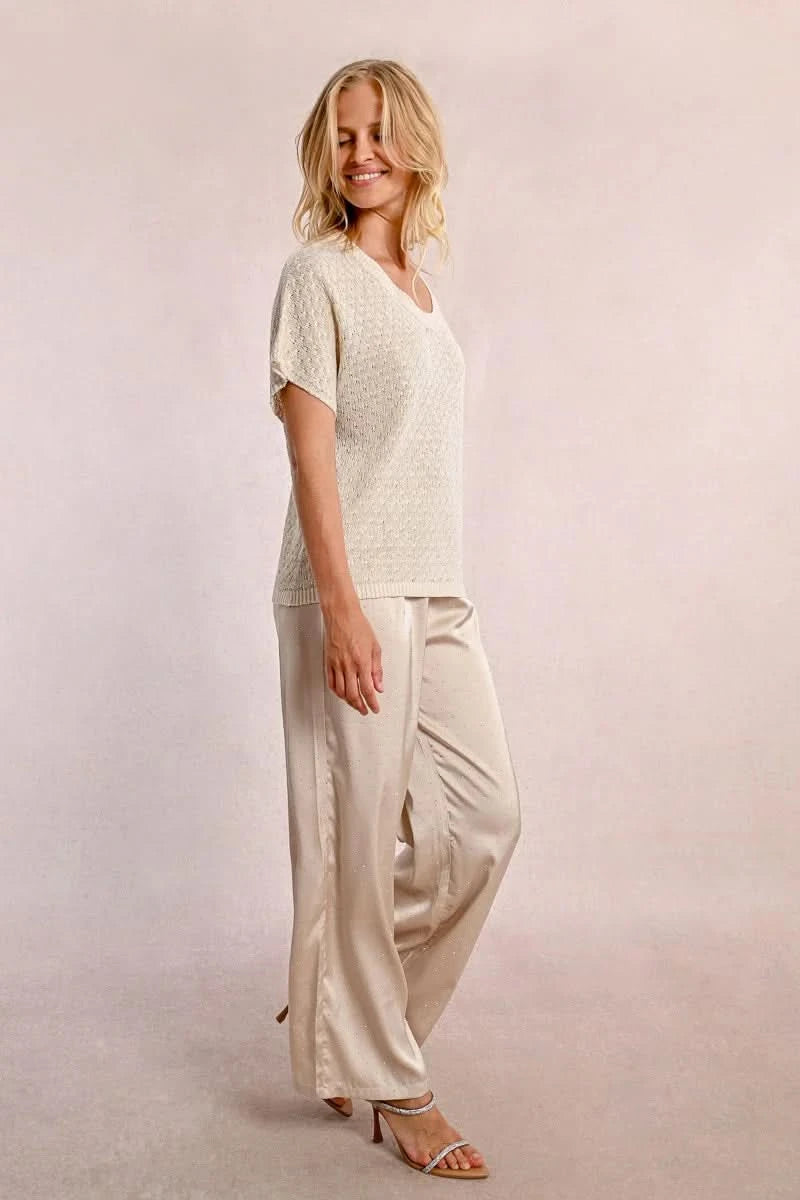 A person with long blond hair stands against a light wall, hands in pockets, wearing the Short Sleeve Knit Top by Molly Bracken and loose pants.