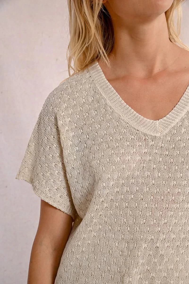 A person with long blond hair stands against a light wall, hands in pockets, wearing the Short Sleeve Knit Top by Molly Bracken and loose pants.