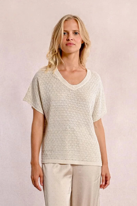 A person with long blond hair stands against a light wall, hands in pockets, wearing the Short Sleeve Knit Top by Molly Bracken and loose pants.