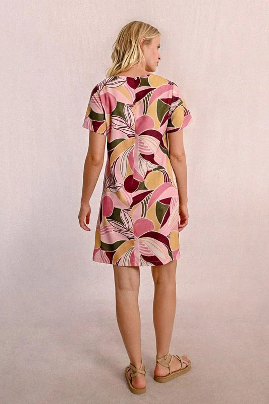 A woman in a Molly Bracken Floral Print T Shirt Dress, featuring a round collar, practical pockets, and short sleeves, stands against a neutral background. Her long blonde hair complements the vibrant outfit she pairs with chic sandals.