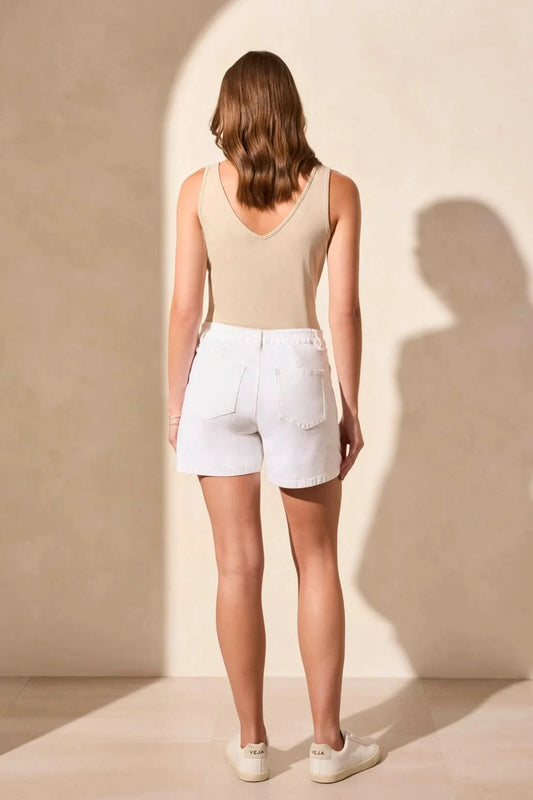 A woman stands against a light-colored wall, wearing a beige sleeveless top and white Tribal PLEATED WIDE SHORTS with back elastic and pockets, her hands behind her back.