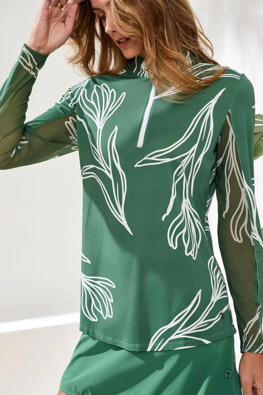 A woman stands indoors on a light woven rug, wearing Tribal's PERFORMANCE QUARTER-ZIP LONG SLEEVE in green with white leaf patterns, a matching skirt, white socks, and beige sneakers.