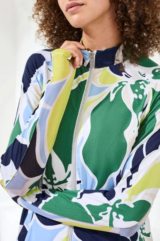 A woman wears the Tribal HIGH PERFORMANCE MESH MOCK-NECK ZIP-UP dress with a blue, green, and white leaf pattern and UPF 50+ protection, standing indoors with her hand in a pocket.