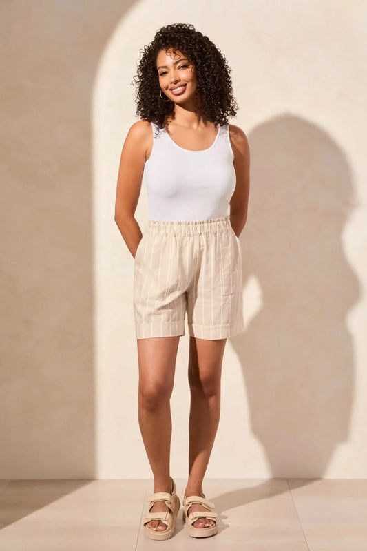 A woman with curly hair stands against a neutral wall, wearing Tribal’s PULL ON SHORT WITH RUFFLE WAISTBAND in beige stripes, a white tank top, and beige sandals—her hands tucked in her pockets for a relaxed summer look.