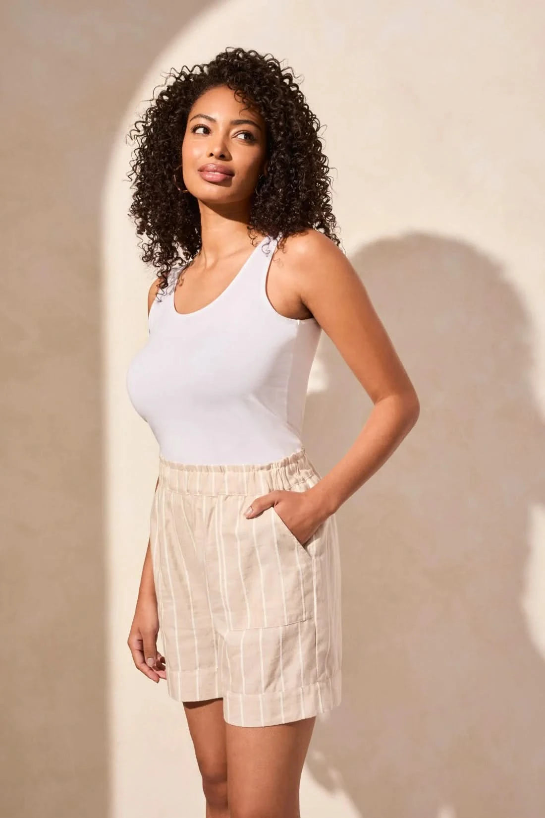 A woman with curly hair stands against a neutral wall, wearing Tribal’s PULL ON SHORT WITH RUFFLE WAISTBAND in beige stripes, a white tank top, and beige sandals—her hands tucked in her pockets for a relaxed summer look.