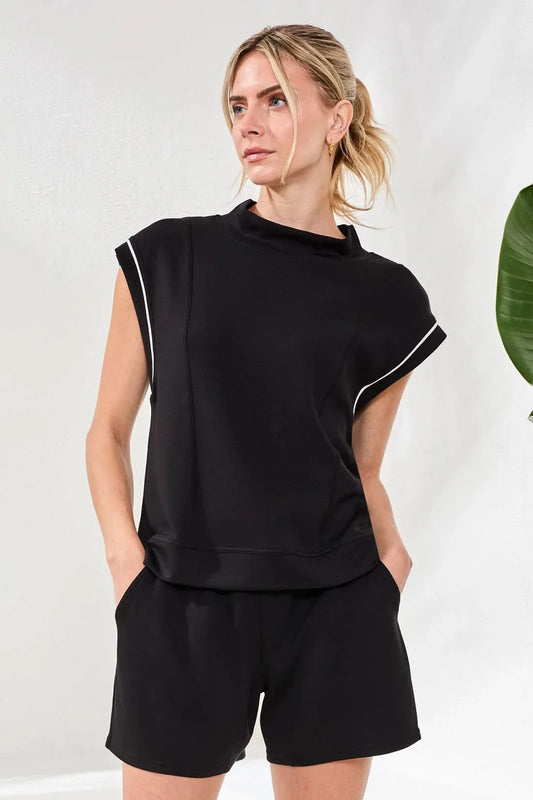 A woman poses indoors in athletic style wearing Tribal's TECHNO LUX MOCK NECK CONTRAST PIPING top and matching shorts with white sneakers and socks, hands in pockets, beside a potted plant.