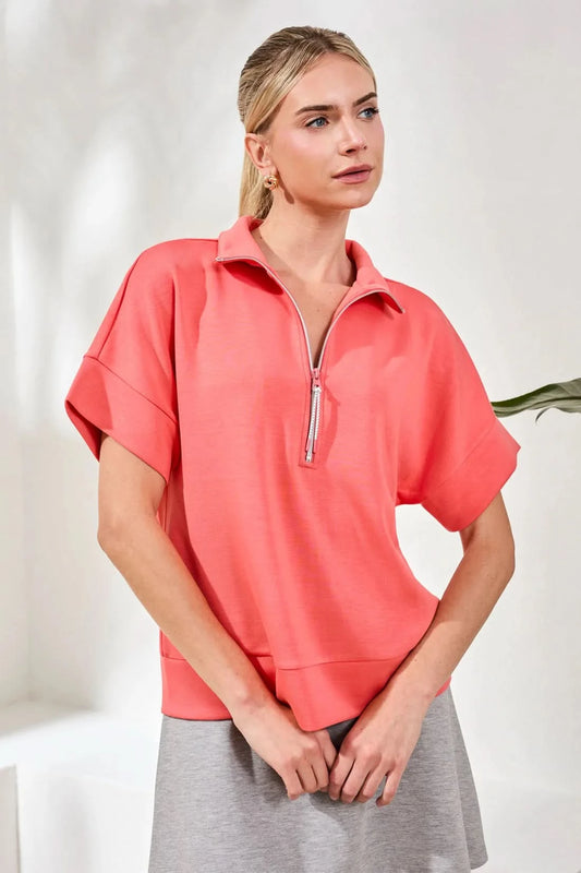 A woman stands indoors next to a green plant wearing the Tribal TECHNO LUX MOCK NECK coral short-sleeve top with zip detail, a light gray skirt, white socks, and sneakers on a woven rug.