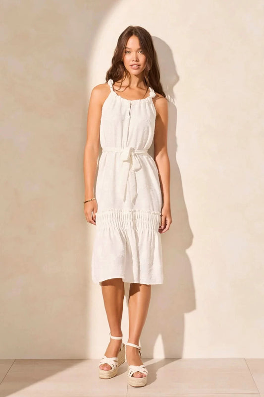 A woman stands in a sunlit room, stylishly cool in the Tribal LINED DRESS WITH PLEAT DETAIL, a sleeveless white summer style paired with wedge sandals as she looks over her shoulder.