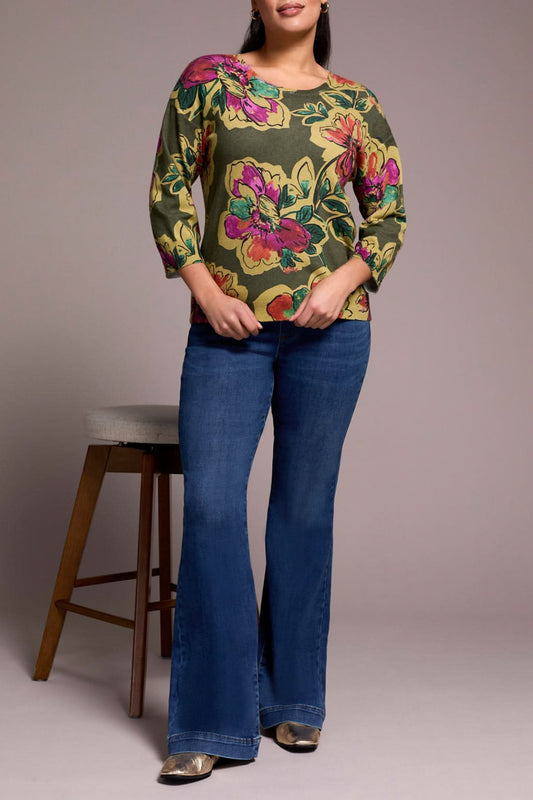 A woman stands beside a wooden stool, wearing the Tribal 3/4 sleeve sweater in Chive and blue flared jeans with silver shoes.