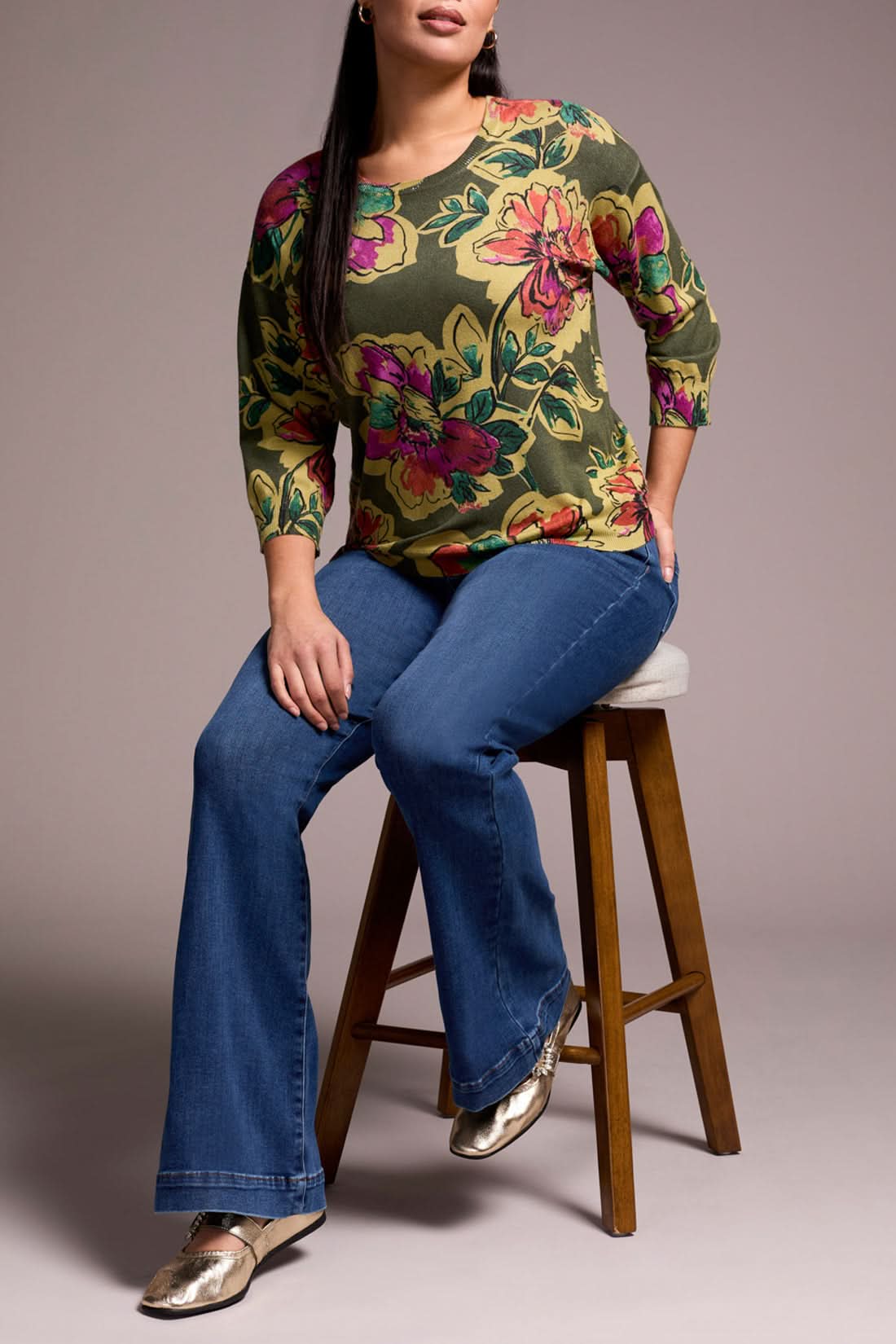 A woman stands beside a wooden stool, wearing the Tribal 3/4 sleeve sweater in Chive and blue flared jeans with silver shoes.