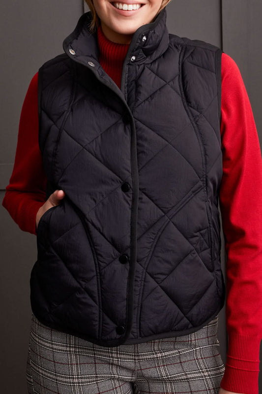 Person wearing a cozy black A Line Puffer Vest from Tribal over a red sweater with plaid pants, making a fashion statement while standing in front of a gray background.