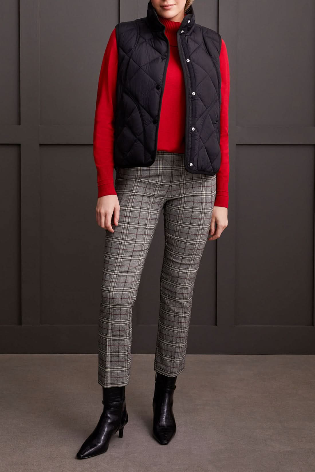 Person wearing a cozy black A Line Puffer Vest from Tribal over a red sweater with plaid pants, making a fashion statement while standing in front of a gray background.