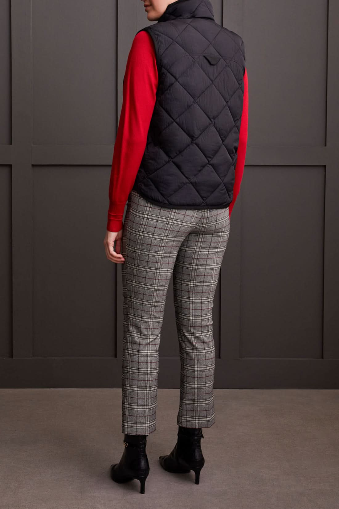 Person wearing a cozy black A Line Puffer Vest from Tribal over a red sweater with plaid pants, making a fashion statement while standing in front of a gray background.