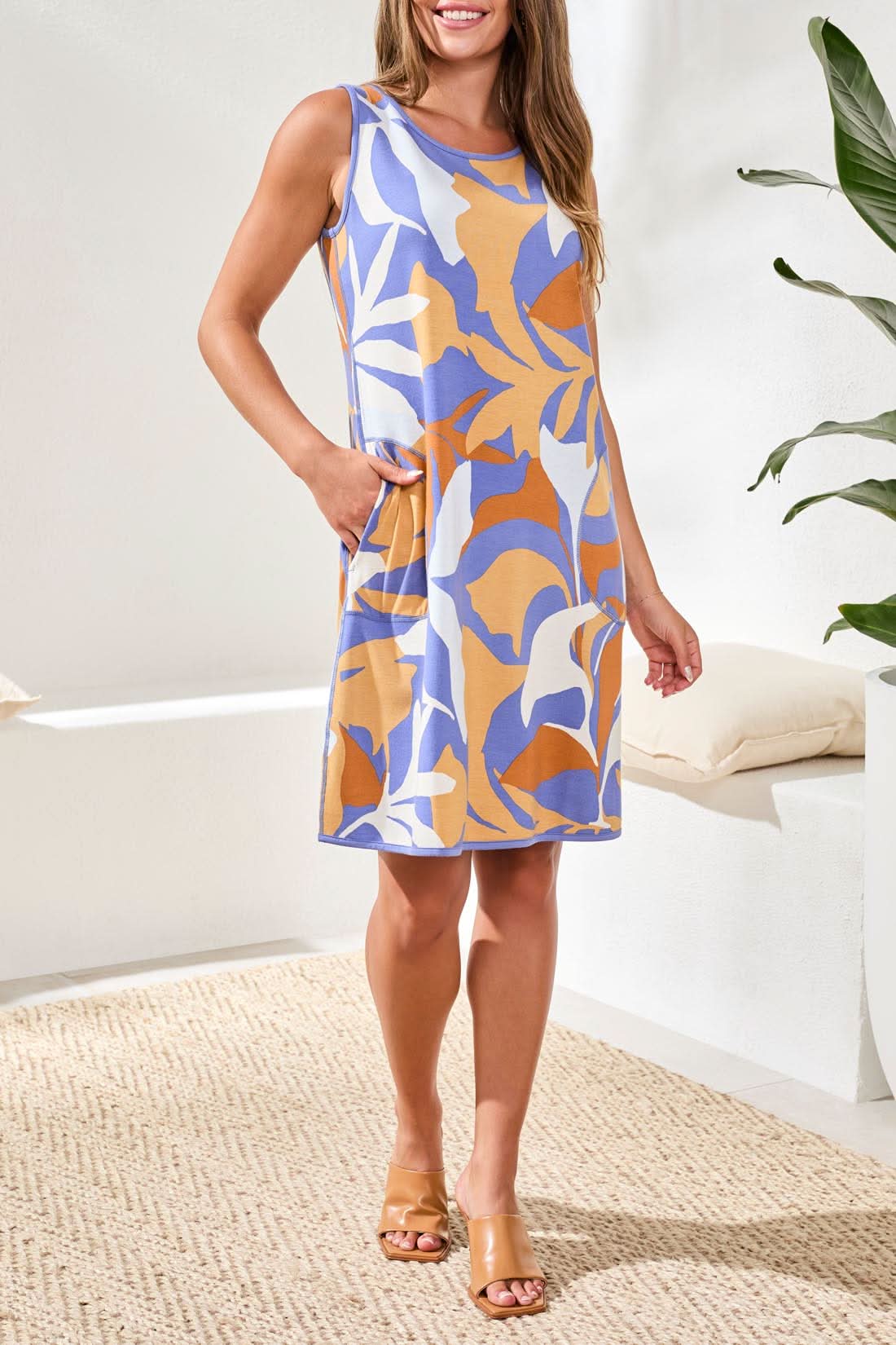 A woman stands indoors on a woven rug by a plant, wearing the Tribal Short Sleeve Summer Dress featuring a blue, white, and orange abstract print.