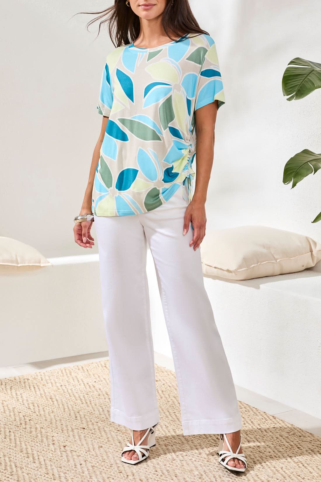A woman wearing a Tribal Boat Neck Top in aqua sky with side ruching and white pants stands indoors near a cushion.