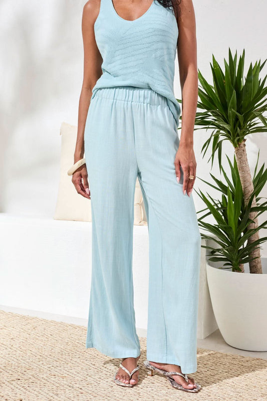 A woman models the Tribal Wide Leg Breezy Pant in light blue, paired with a matching sleeveless top, standing indoors on a woven rug near green plants and a white bench with beige cushions.