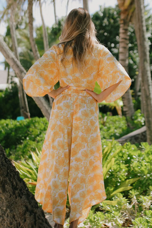 A woman with long hair stands outdoors among lush greenery, wearing Tribal's PRINTED COVER-UP PANTS WITH SASH in a yellow and white floral pattern—perfect for a summer getaway.
