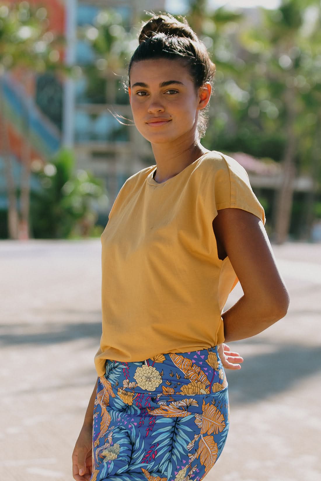 A woman stands outdoors in the Tribal CAP SLEEVE CREW NECK TOP and a blue floral skirt, with palm trees and a building behind her—a stylish choice for your next vacation wardrobe.