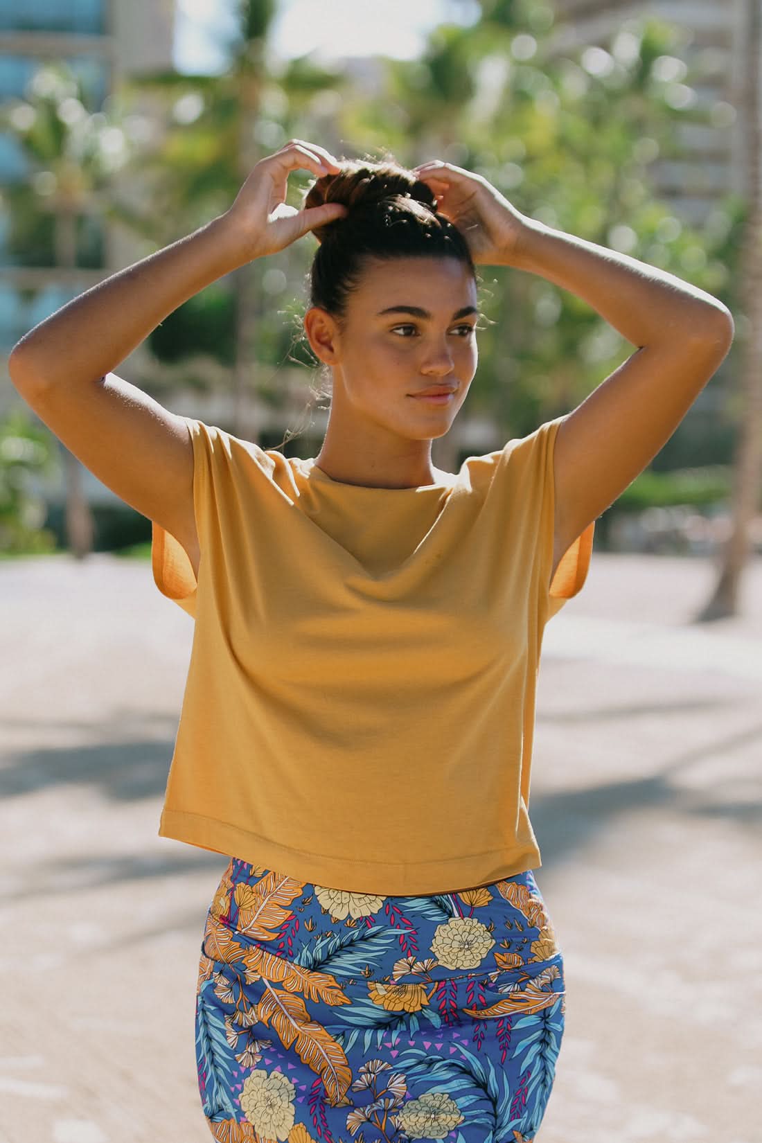 A woman stands outdoors in the Tribal CAP SLEEVE CREW NECK TOP and a blue floral skirt, with palm trees and a building behind her—a stylish choice for your next vacation wardrobe.