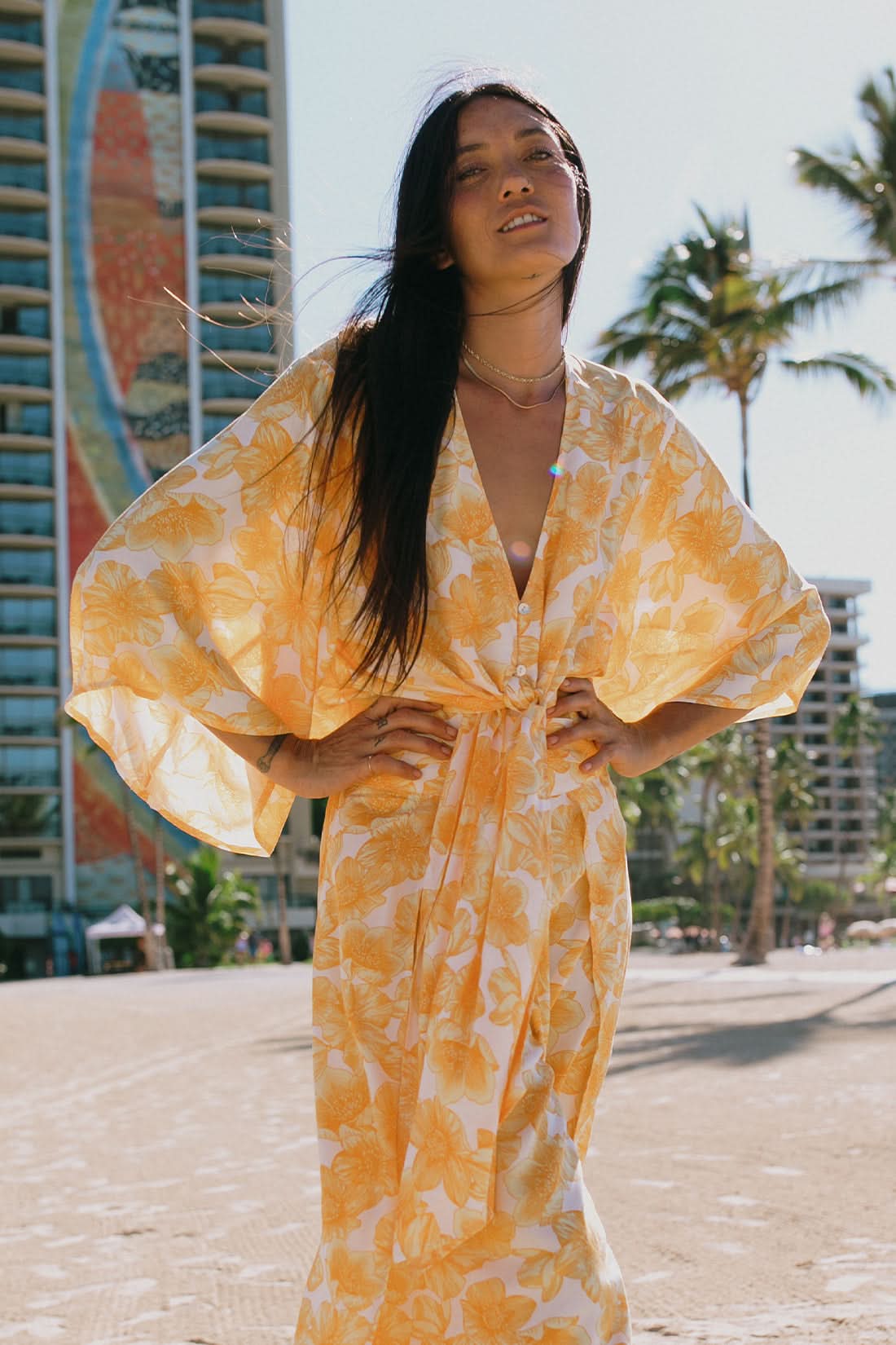 A woman stands outdoors near tropical greenery and palm trees wearing the Tribal KIMONO TOP WITH FRONT TIE, a yellow and white floral two-piece outfit featuring kimono-style sleeves.