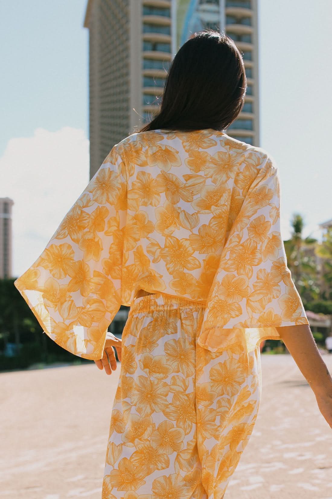 A woman stands outdoors near tropical greenery and palm trees wearing the Tribal KIMONO TOP WITH FRONT TIE, a yellow and white floral two-piece outfit featuring kimono-style sleeves.