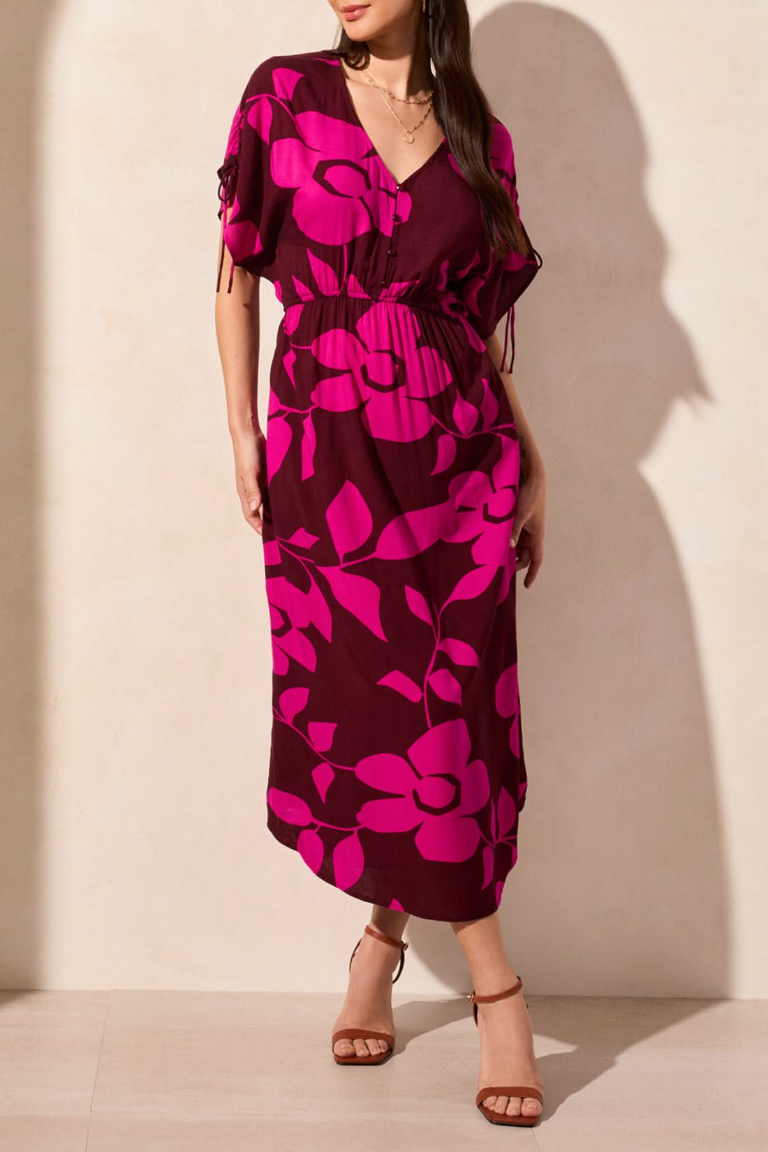 A woman wears the Tribal Flowy Dress with Shoulder Ties—featuring bright pink floral patterns, a V-neckline, and short sleeves—paired with brown strappy heels against a beige wall, creating an effortless summer look.
