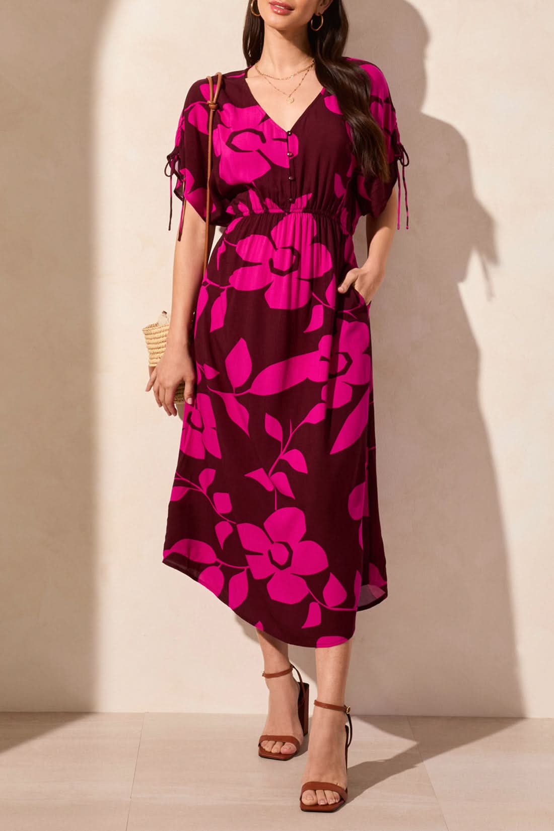 A woman wears the Tribal Flowy Dress with Shoulder Ties—featuring bright pink floral patterns, a V-neckline, and short sleeves—paired with brown strappy heels against a beige wall, creating an effortless summer look.