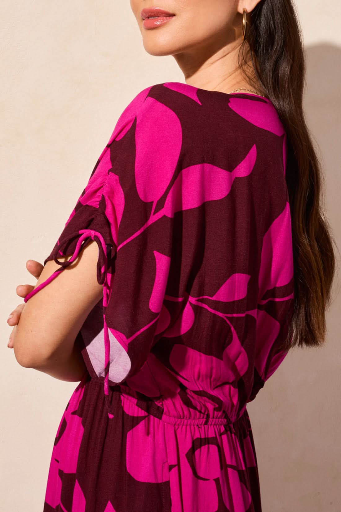 A woman wears the Tribal Flowy Dress with Shoulder Ties—featuring bright pink floral patterns, a V-neckline, and short sleeves—paired with brown strappy heels against a beige wall, creating an effortless summer look.