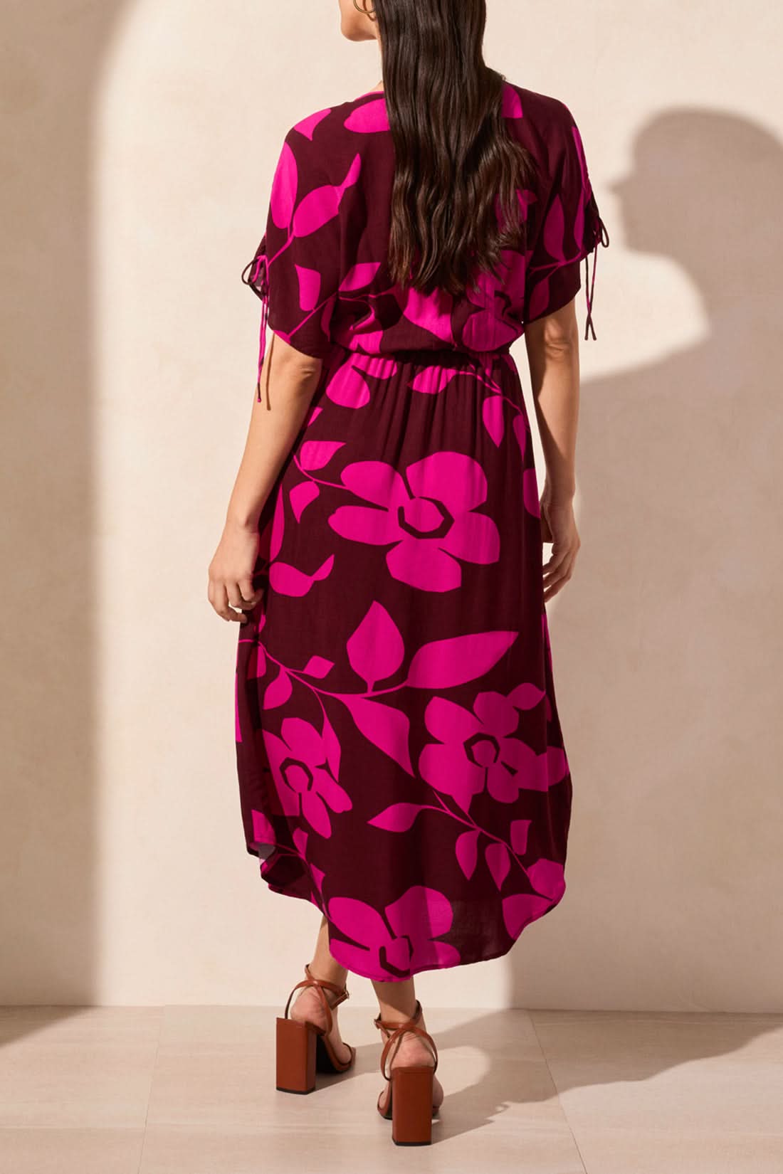 A woman wears the Tribal Flowy Dress with Shoulder Ties—featuring bright pink floral patterns, a V-neckline, and short sleeves—paired with brown strappy heels against a beige wall, creating an effortless summer look.
