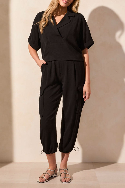 A person stands indoors facing away, wearing the Tribal FLOWY POPOVER SHORT SLEEVE SHIRT for breathable comfort, paired with black cargo pants featuring pockets and drawstring hems, and silver flat sandals.