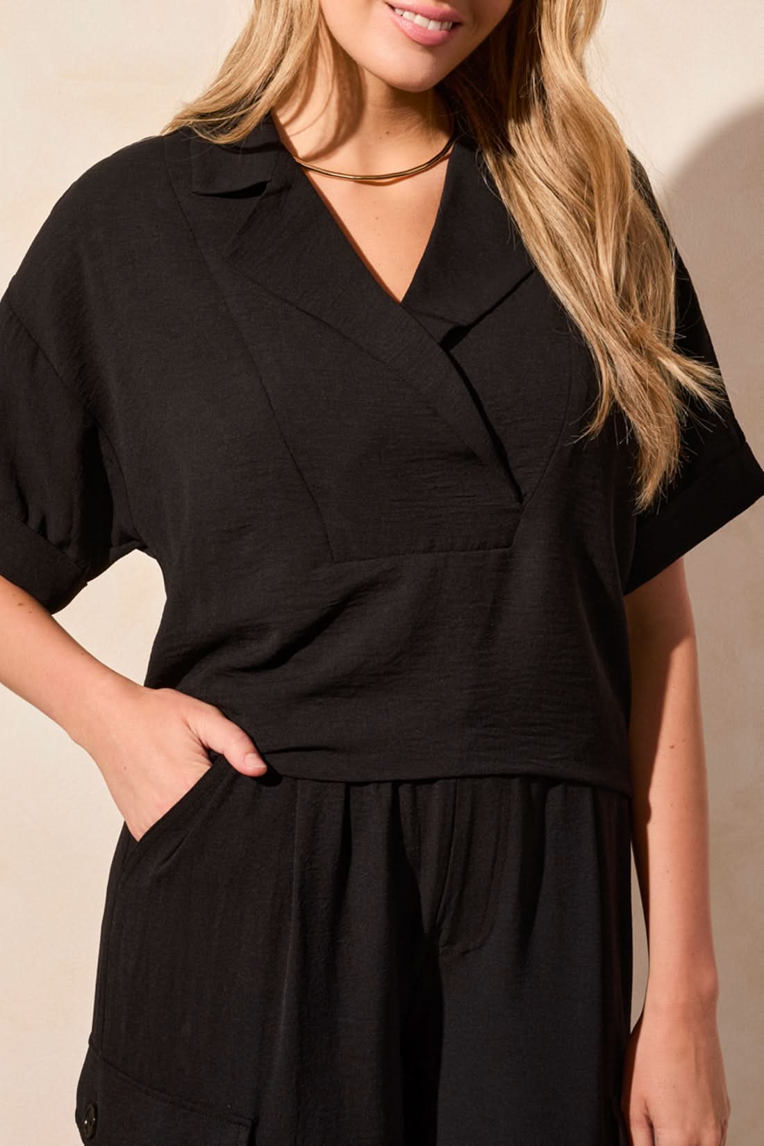 A person stands indoors facing away, wearing the Tribal FLOWY POPOVER SHORT SLEEVE SHIRT for breathable comfort, paired with black cargo pants featuring pockets and drawstring hems, and silver flat sandals.