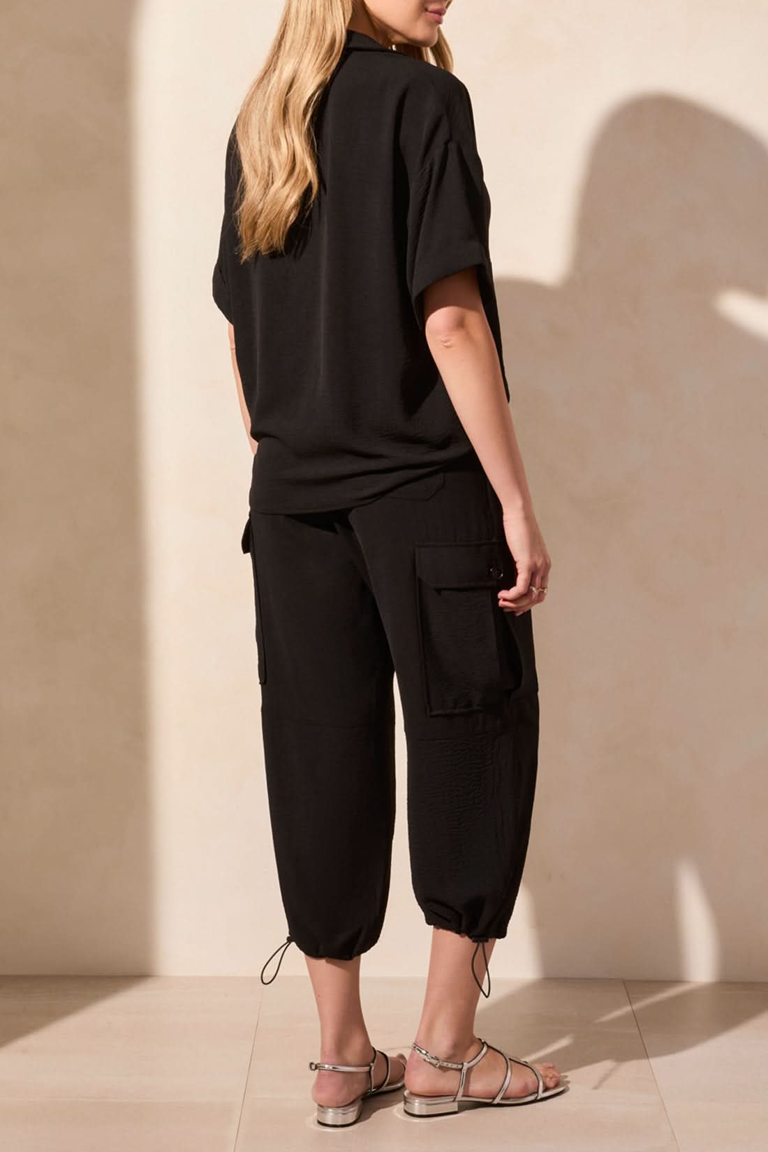 A woman wearing the Tribal Flowy Popover Short Sleeve Shirt in black, styled with matching black pants, stands with one hand in her pocket against a beige background.