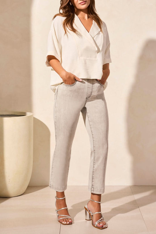 A woman stands indoors, facing away, wearing the Tribal FLOWY POPOVER SHORT SLEEVE SHIRT in white, light gray jeans, and metallic block-heeled sandals.